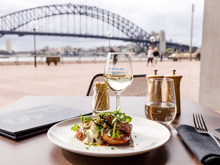 A meal with a glass of wine on a table with the Harbour Bridge in the background
